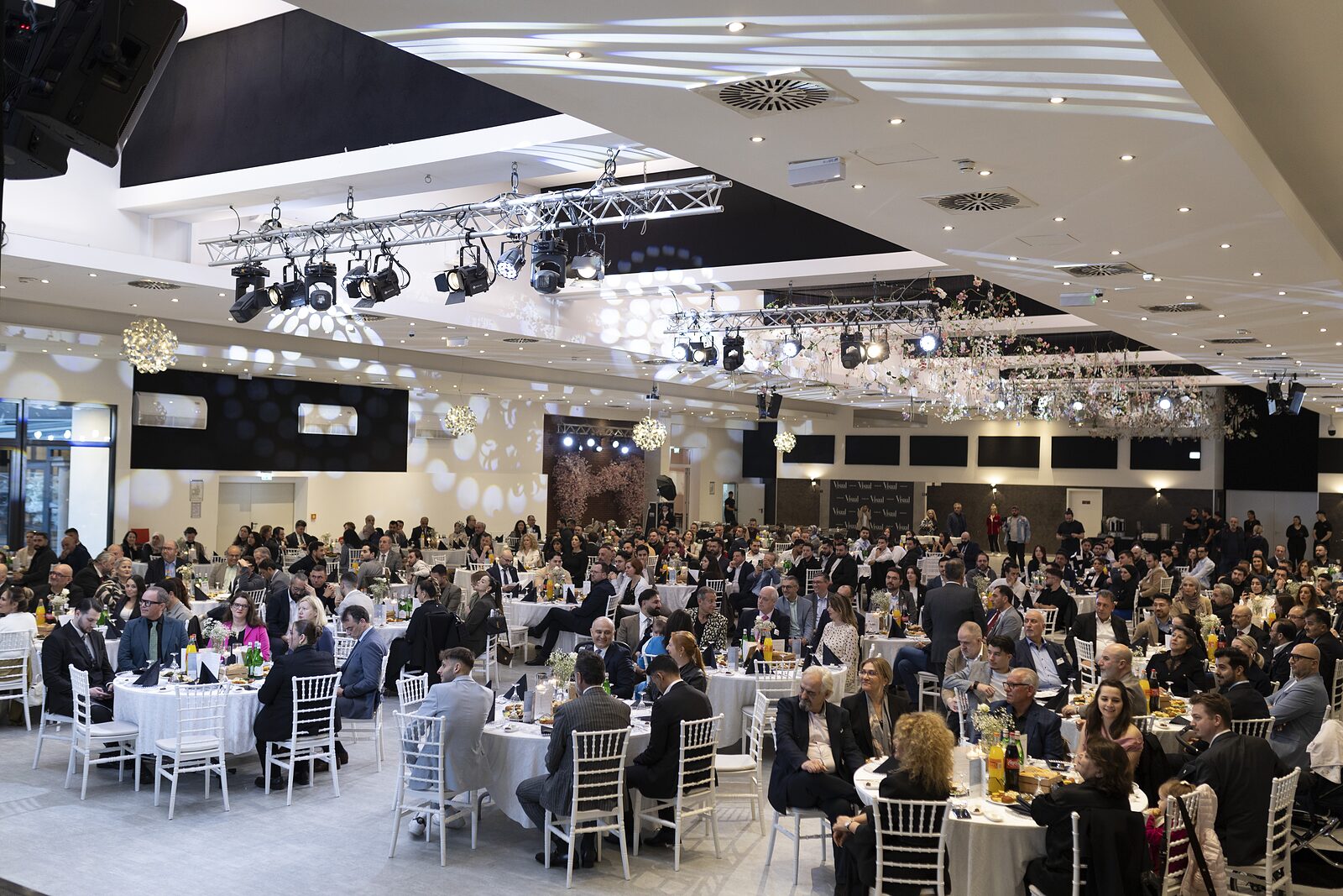 Vollbesetzter großer Saal bei einem Gala-Abend – runde Tafeln, Traversenlicht und Bühne